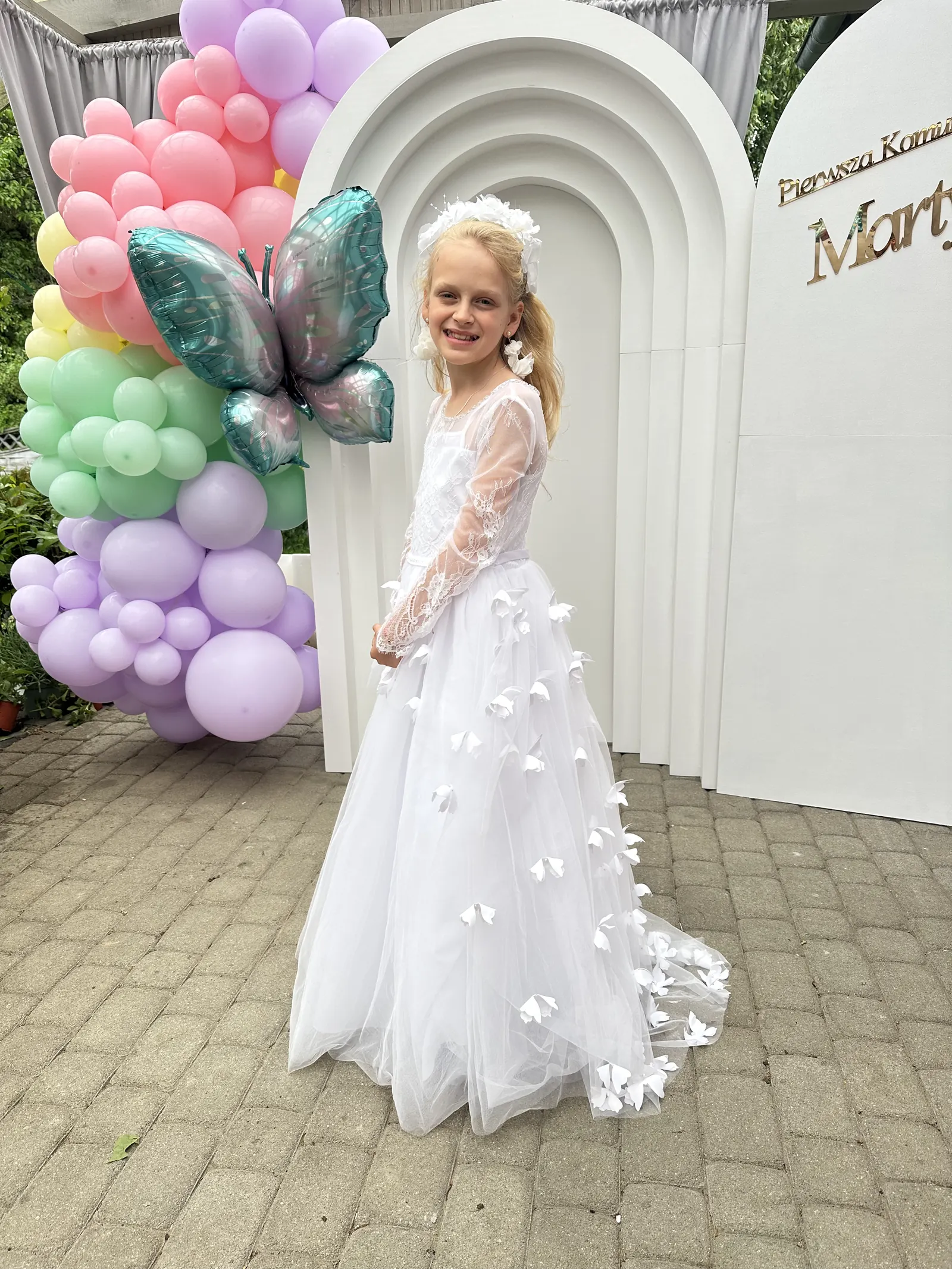 Dziewczynka w sukience komunijnej przy pastelowym łuku balonowym